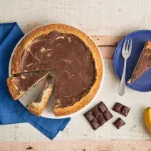 Fotografia em tons de azul em uma bancada de madeira, um pano azul, um prato grande raso com a cheesecake de banana com chocolate em cima dele. Ao lado, um prato pequeno azul raso com uma fatia da cheesecake, um cacho de banana e quadradinhos de chocolate Fotografia em tons de azul em uma bancada de madeira, um pano azul, um prato grande raso com a cheesecake de banana com chocolate em cima dele. Ao lado, um prato pequeno azul raso com uma fatia da cheesecake, um cacho de banana e quadradinhos de chocolate